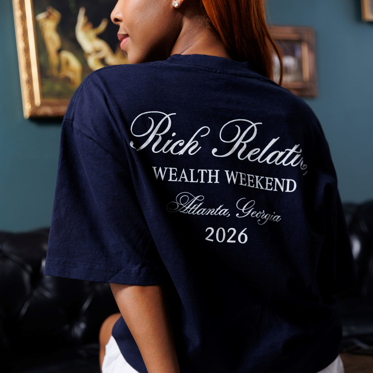 A woman wearing a navy blue oversized T-shirt with white script text reading 'Rich Relatives Wealth Weekend Atlanta, Georgia 2026' on the back, seated on a wooden trunk in an indoor setting.
