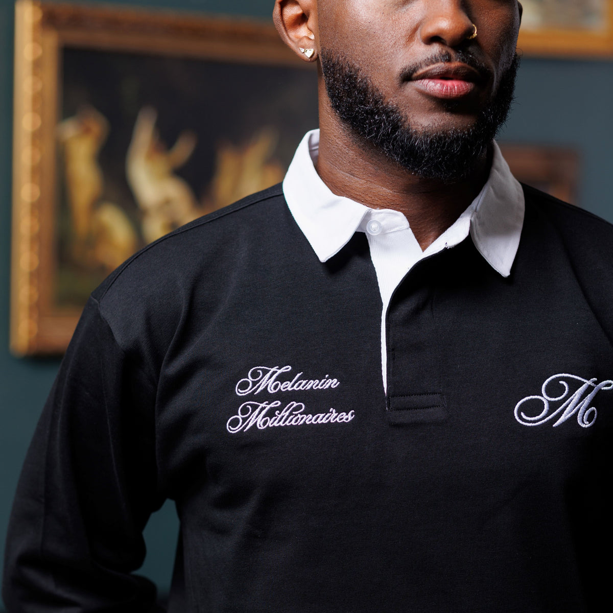 A man wearing a black long-sleeve rugby-style polo shirt with a white collar. The shirt features white embroidered script logos on the chest, one reading 'Melanin Modernaires' and the other a stylized 'M'.